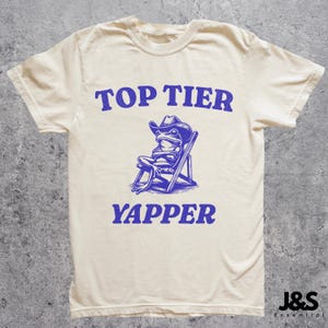 以下が含まれることがあります： クリーム色のTシャツに、カウボーイハットをかぶって椅子に座っているカエルの青いグラフィックが描かれています。Tシャツには「TOP TIER YAPPER」という青い文字がプリントされています。