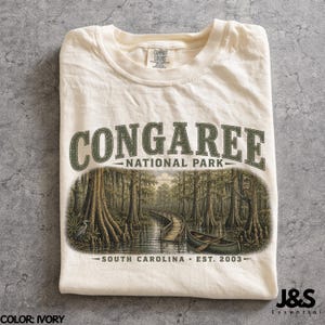 May include: Ivory-colored t-shirt featuring a graphic of Congaree National Park in South Carolina. The design includes the text "CONGAREE NATIONAL PARK" above a detailed illustration of a swamp scene with a canoe and boardwalk. The text "SOUTH CAROLINA EST. 2003" is also included.