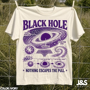 Op de afbeelding: Een ivoorkleurig t-shirt met een paarse afbeelding van een zwart gat, gelabeld met event horizon, accretion disk en singularity. Daaronder staat een diagram in drie stappen met de tekst PULL! en NOTHING ESCAPES THE PULL.