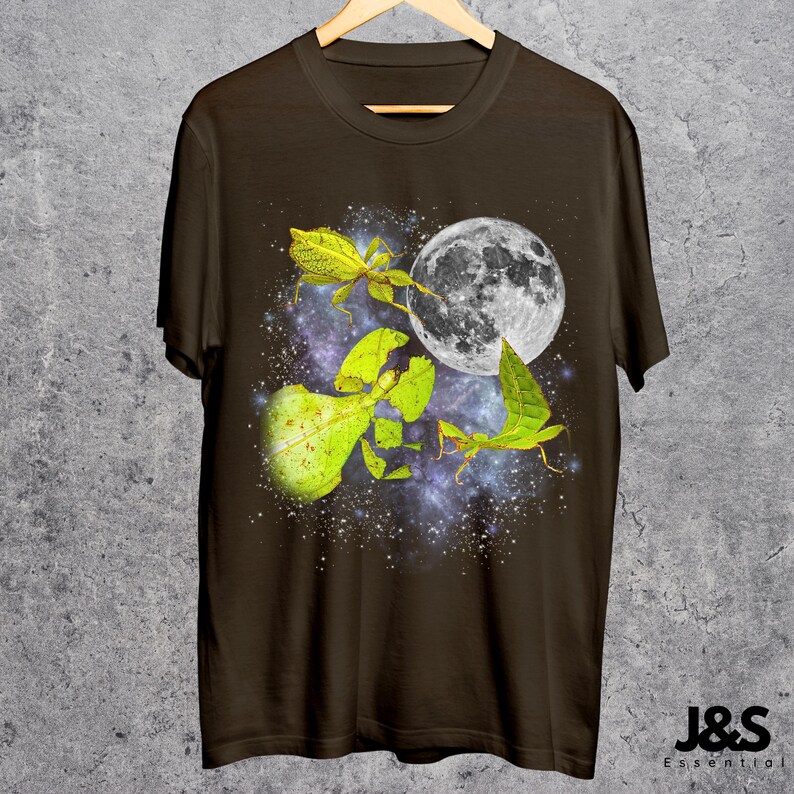 May include: Brown t-shirt featuring a vibrant graphic of leaf insects with a full moon against a starry galaxy backdrop. The design includes detailed illustrations of the insects in shades of green and yellow. The shirt has the text "J&S Essential" at the bottom.