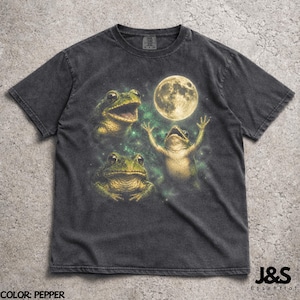 Può includere: T-shirt color pepe con un design di tre rane e una luna piena. Le rane sono raffigurate in varie pose, con una che si protende verso la luna. La maglietta ha un aspetto vintage. Il testo "J&S Essential" è in basso.