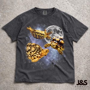 May include: Dark gray t-shirt featuring a graphic of three tortoises in space with a full moon and starry background. The tortoises are yellow and brown. The shirt has a vintage wash look and the text "J&S Essential" is at the bottom.