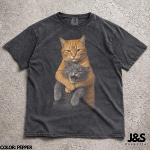 May include: Pepper-coloured t-shirt with a graphic of two cats. An orange cat hugs a grey cat, which has its mouth open. The text "J&S Essential" is in the lower right corner. The colour of the shirt is "Pepper".