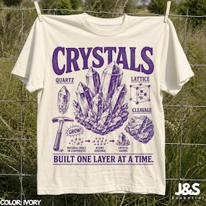 May include: An ivory t-shirt featuring a purple graphic print of various crystals, a hammer, and scientific diagrams. The text reads "CRYSTALS," "QUARTZ," "LATTICE," "CLEAVAGE," "GROW!," "MATERIAL COOLS OR EVAPORATES," "ATOMS ARRANGE," "CRYSTAL GROWS," and "BUILT ONE LAYER AT A TIME."