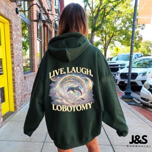 May include: Dark green hoodie with the words "LIVE, LAUGH, LOBOTOMY" above a graphic of a dolphin jumping through a colorful, celestial design. The hoodie is a solid color with a drawstring hood.
