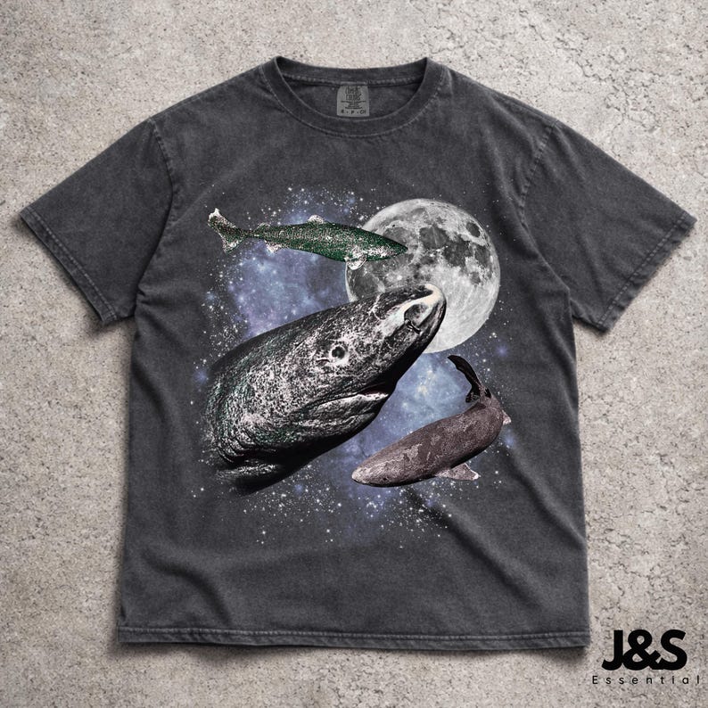 以下が含まれることがあります： 魚、満月、星空のシュールなグラフィックが特徴の濃いグレーのTシャツ。デザインには大きな魚の頭と他の2匹の魚が含まれています。シャツはヴィンテージ感があり、下部に「J&S Essential」の文字があります。