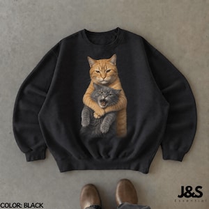 Può includere: Una felpa nera a girocollo con una stampa grafica di un gatto soriano arancione che abbraccia un gatto grigio più piccolo. Il gatto arancione ha un'espressione calma, mentre il gatto grigio ha la bocca aperta. Testo J&S Essential presente.