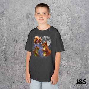 May include: A dark gray t-shirt featuring a whimsical design of three squirrels against a backdrop of a full moon and a starry sky. The squirrels are depicted in shades of brown and orange. The shirt has the text "J&S Essential" at the bottom.