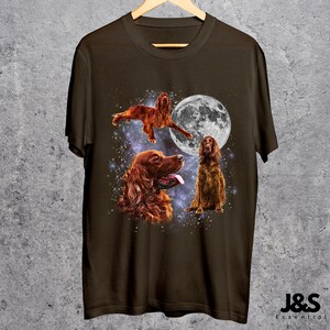 K&ouml;nnte beinhalten: Dunkelbraunes T-Shirt mit Illustrationen von Irish Setter Hunden, einem Vollmond und einem Sternenhimmel. Das Design zeigt drei Hunde in verschiedenen Posen. Das T-Shirt h&auml;ngt an einem Holzkleiderb&uuml;gel.