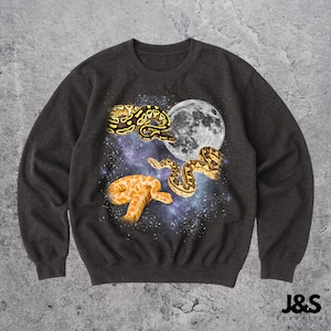 Peut inclure: Sweat-shirt gris foncé avec un motif céleste. Il présente trois serpents, une pleine lune et un fond de galaxie étoilée. Les serpents sont jaunes et bruns. Le sweat-shirt porte le texte "J&S Essential" en bas à droite.