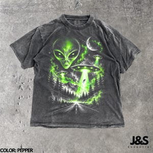 Könnte beinhalten: Ein anthrazitfarbenes Kurzarm-T-Shirt mit einem Grafikdruck eines großen grünen Alien-Kopfes, einer Mondsichel und mehreren fliegenden Untertassen, die hellgrüne Strahlen über eine dunkle Waldlandschaft und eine Straße in die Ferne aussenden.
