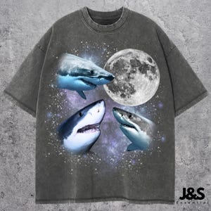 May include: A gray t-shirt with a graphic print of three white sharks and a full moon. The sharks are swimming in a galaxy-like background.