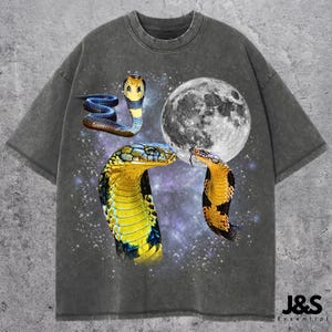 May include: Grey t-shirt with a graphic of three cobras, a full moon, and a starry backdrop. The snakes are yellow, blue, and black. The text "J&S Essential" is at the bottom right.