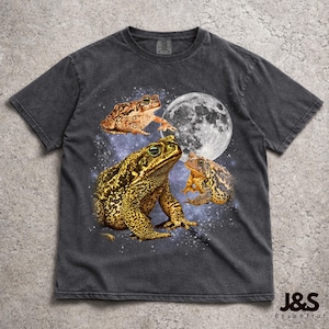 Puede incluir: Camiseta gris oscuro con un diseño de tres ranas, una luna llena y un fondo estrellado. Las ranas son marrones y doradas. La camiseta tiene un aspecto vintage y desgastado.