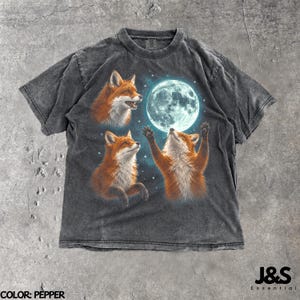 Peut inclure: Un t-shirt gris anthracite délavé à l'acide avec une impression graphique représentant trois renards orange et une pleine lune brillante entourée de petites étoiles blanches. Le texte J&S Essential et Color: Pepper sont visibles.