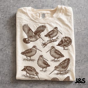 May include: Cream-colored t-shirt featuring detailed sepia-toned illustrations of various birds, including one in flight and several perched or standing. The shirt is folded neatly, with the brand label visible at the neckline. The lower right corner displays the text "J&S Essential".