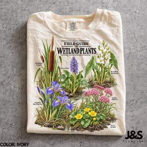 May include: An ivory t-shirt featuring a graphic titled Field Guide to Wetland Plants. The print displays illustrations of Broadleaf Cattail, Pickerelweed, Broadleaf Arrowhead, Northern Blue Flag Iris, Swamp Milkweed, and Marsh Marigold with labels.