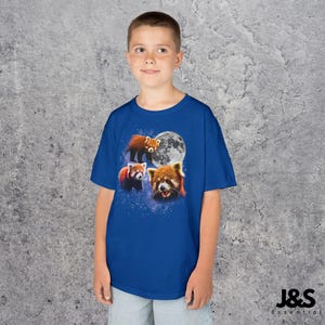 May include: A blue t-shirt featuring a design of three red pandas with a full moon in the background. The red pandas are in different poses, with one standing, one sitting, and one with its mouth open. The shirt has the text "J&S Essential" at the bottom.