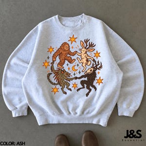 May include: An ash gray crewneck sweatshirt featuring a central graphic of four mythical creatures dancing in a circle. The design includes a brown sasquatch, a jackalope, a small green creature, and a dark horned figure, surrounded by orange stars and a crescent moon.