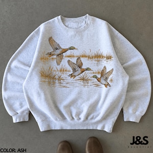 May include: An ash gray crewneck sweatshirt featuring a graphic print of three mallard ducks in flight over a marshy landscape with tall golden grasses and water. The garment has long sleeves, a ribbed collar, and a ribbed hemline.