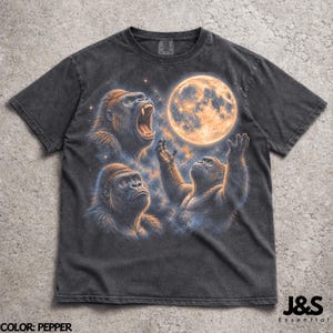 Puede incluir: Camiseta color pimienta con un gráfico de tres gorilas mirando una luna llena. Un gorila ruge, otro mira hacia arriba y el tercero se extiende hacia la luna. La camiseta tiene un lavado vintage.