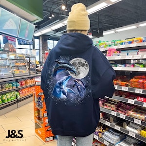 Puede incluir: Sudadera con capucha azul marino con un gráfico de tres delfines saltando frente a una luna llena y un fondo estrellado. La sudadera tiene una capucha con cordón y un bolsillo de canguro. El logotipo de la marca "J&S Essential" está en la parte inferior.