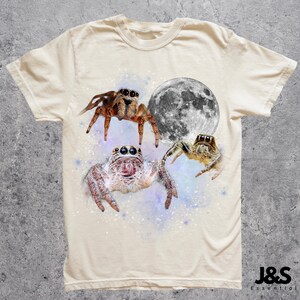 May include: Cream-colored t-shirt featuring a graphic design of three jumping spiders and a full moon. The spiders are brown, white, and tan, set against a starry, purple and blue background. The shirt has a crew neck and short sleeves.