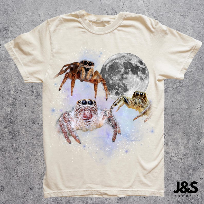 May include: Cream-colored t-shirt featuring a graphic design of three jumping spiders and a full moon. The spiders are brown, white, and tan, set against a starry, purple and blue background. The shirt has a crew neck and short sleeves.