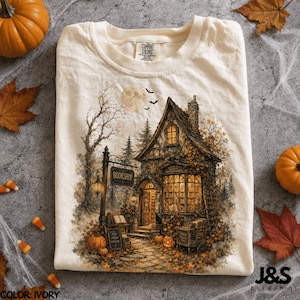 May include: An ivory t-shirt featuring a graphic illustration of a multi-story bookshop with a lit interior, a hanging sign that reads BOOKSHOP, a full moon, flying bats, and pumpkins scattered on the ground amidst fallen autumn leaves.