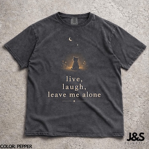 May include: Pepper-colored t-shirt featuring a cat silhouette under a crescent moon and stars. The shirt has the text "live, laugh, leave me alone" in a serif font. The shirt is a crew neck and has short sleeves.