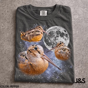Op de afbeelding: Een antracietgrijs t-shirt met een grafische print van drie bruine houtsnippen tegen een achtergrond van een volle maan en een sterrenstelsel. Het t-shirt heeft een Comfort Colors merklabel in de nek en de tekst J&S Essential onderaan.