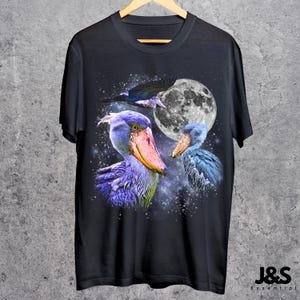 Puede incluir: Camiseta negra con una ilustraci&oacute;n detallada de tres aves picozapato sobre un cielo nocturno con luna llena. Las aves est&aacute;n representadas con plumaje azul, morado y naranja. La camiseta incluye el texto "J&S Essential" en la parte inferior.