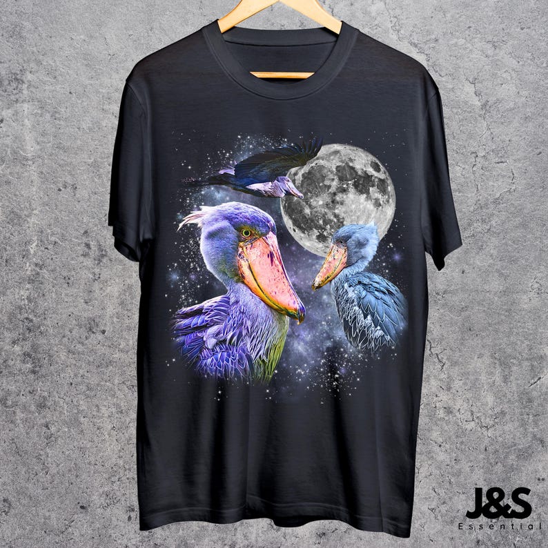 Puede incluir: Camiseta negra con una ilustraci&oacute;n detallada de tres aves picozapato sobre un cielo nocturno con luna llena. Las aves est&aacute;n representadas con plumaje azul, morado y naranja. La camiseta incluye el texto "J&S Essential" en la parte inferior.