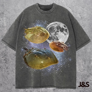Puede incluir: Camiseta gris oscuro con un diseño gráfico de tres cangrejos herradura, una luna llena y un fondo estrellado. La camiseta tiene un efecto de lavado vintage y el texto "J&S Essential" en la parte inferior.