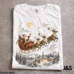Pode incluir: Uma t-shirt branca com uma estampa gráfica do Pai Natal num trenó vermelho puxado por três renas a voar sobre uma paisagem de aldeia nevada com uma lua cheia no céu. A t-shirt tem um decote redondo e uma etiqueta Comfort Colors.