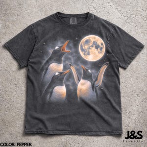 Könnte beinhalten: Pfefferfarbenes T-Shirt mit einer Grafik von drei Pinguinen, die unter einem Vollmond singen. Die Pinguine haben orangefarbene Schnäbel und Brustbereiche, mit einem sternenklaren Nachthimmel im Hintergrund. Der Text "J&S Essential" befindet sich unten rechts.