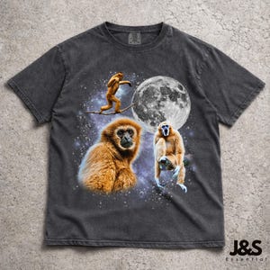 Puede incluir: Camiseta gris oscuro con un diseño de tres gibones sobre un cielo estrellado y una luna llena. Los gibones están representados en tonos marrones y beige. El texto "J&S Essential" está en la parte inferior derecha.