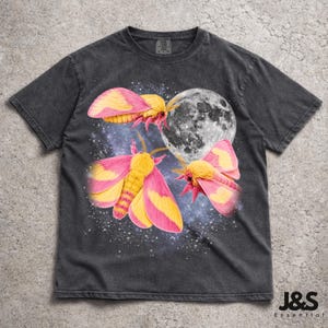 May include: A charcoal gray short-sleeve t-shirt featuring a graphic print of three vibrant pink and yellow moths surrounding a detailed white moon against a starry, dark blue cosmic background. The shirt has a crew neckline and a relaxed fit.