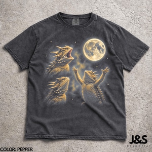 May include: Pepper-colored t-shirt featuring a graphic of three horned lizards with a full moon in the background. One lizard is raising its arms, another is roaring, and the third is looking up. The text "J&S Essential" is in the lower right corner.