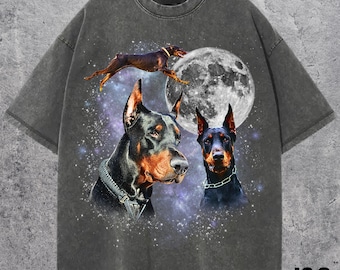 Puede incluir: Camiseta gris oscuro con un gráfico de tres Doberman Pinschers, una luna llena y un fondo estrellado. Un perro salta, mientras que los otros dos miran hacia adelante. El texto "J&S Essential" está en la parte inferior derecha.