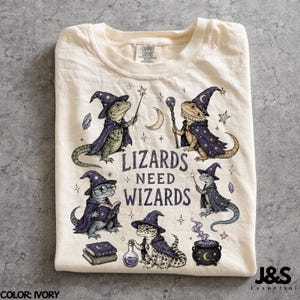 May include: An ivory t-shirt featuring a graphic print of five lizards dressed as wizards in purple capes and pointed hats. The design includes stars, a crescent moon, books, a potion bottle, a cauldron, and the text "LIZARDS NEED WIZARDS".