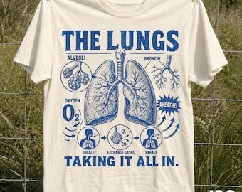 Könnte beinhalten: Ein elfenbeinfarbenes T-Shirt mit einem blauen Grafikdruck menschlicher Lungen und anatomischen Beschriftungen wie Alveoli, Bronchi und Oxygen. Das Design zeigt Diagramme des Ein- und Ausatmungsprozesses mit dem Text THE LUNGS TAKING IT ALL IN.