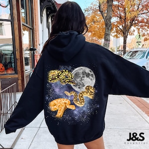 Könnte beinhalten: Schwarzer Kapuzenpullover mit einem grafischen Design mit Schlangen, einem Vollmond und einem Sternenhintergrund. Der Pullover hat den Text "J&S Essential" unten rechts.