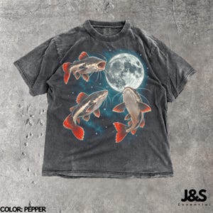 Op de afbeelding: Een antracietgrijs T-shirt met korte mouwen en een grafische print van drie meervallen die rond een grote, stralende volle maan zwemmen tegen een donkere, sterrenhemel. Het shirt heeft een verwassen textuur en een ronde hals.
