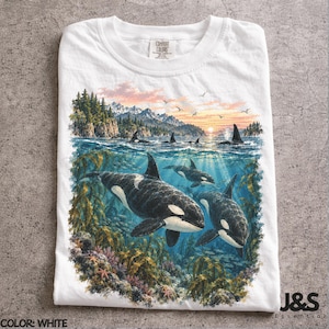 May include: A white t-shirt featuring a detailed graphic print of orca whales swimming underwater among kelp and coral, with a scenic landscape of mountains, pine trees, and a sunset sky visible above the water surface on the front of the garment.