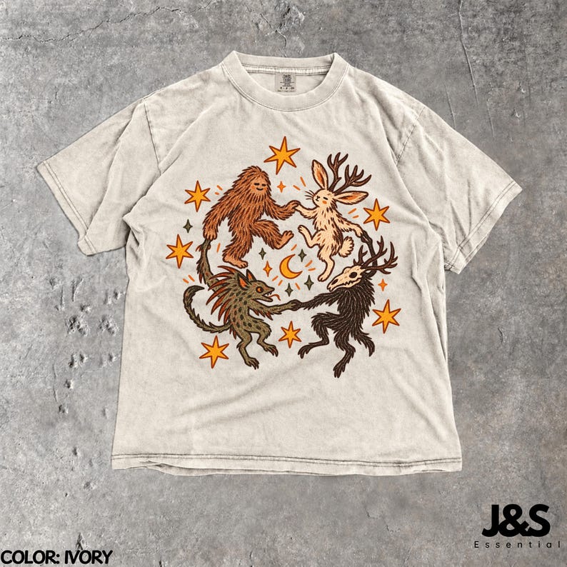May include: An ivory short-sleeve t-shirt featuring a central graphic of a brown sasquatch, a tan jackalope, a green creature, and a dark brown skeletal deer-like figure dancing in a circle surrounded by orange stars and a crescent moon.
