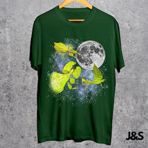 May include: Dark green t-shirt featuring a graphic of a full moon and several leaf insects in shades of yellow and green. The design is set against a starry, galaxy-like background. The shirt is hanging on a wooden hanger.