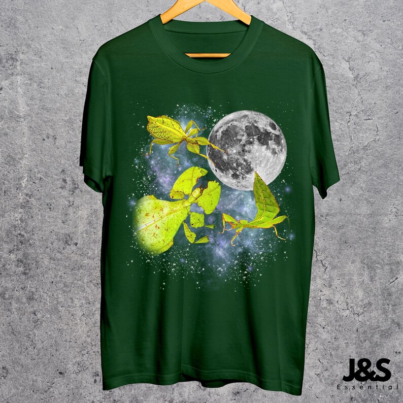 May include: Dark green t-shirt featuring a graphic of a full moon and several leaf insects in shades of yellow and green. The design is set against a starry, galaxy-like background. The shirt is hanging on a wooden hanger.