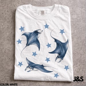 May include: White t-shirt featuring a watercolor design of three manta rays and blue stars. The shirt has a crew neck and is made of a soft material. The text "Combat Colors" is visible on the upper left. The brand "J&S Essential" is on the lower right.