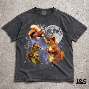 Puede incluir: Camiseta gris oscuro con un diseño caprichoso de tres ardillas bajo una luna llena. Las ardillas están representadas en tonos marrones y naranjas, sobre un fondo estrellado. La camiseta tiene un aspecto vintage lavado y el texto "J&S Essential" está en la parte inferior.
